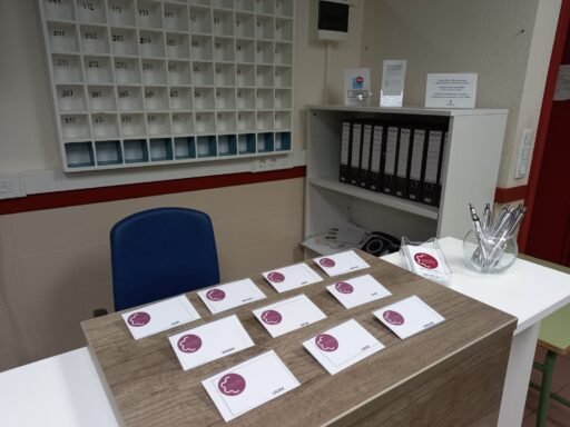 La imagen muestra un mostrador de recepción en una oficina o entorno administrativo. Sobre el mostrador de madera, hay varias etiquetas con un logotipo circular granate que representa el perfil estilizado de un rostro, cada una con nombres individuales como "SARA", "MARIA", "LAURA" y "CAROLINA". A la derecha del escritorio, hay un recipiente de vidrio con bolígrafos plateados y un soporte de plástico transparente con tarjetas adicionales con el mismo logotipo. Detrás del escritorio, hay un sistema de compartimentos de pared con ranuras numeradas, probablemente para correo o distribución de llaves, que van del 101 al 315. También hay una estantería blanca con carpetas etiquetadas y algunos papeles. En la estantería se muestran letreros y tarjetas informativas, pero no se ven marcas específicas ni figuras reconocibles en la imagen. El área parece organizada y lista para el personal o las visitas.