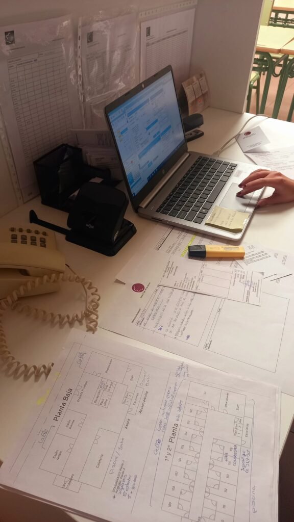 La imagen muestra un espacio de trabajo con una computadora portátil en uso, con una mano visible sobre el teclado. La pantalla muestra lo que parece ser una interfaz de programación o administración con elementos azules. El escritorio está cubierto con diversos documentos y papeles, incluyendo lo que parecen planos de planta etiquetados como "Planta Baja" con la distribución y numeración de las habitaciones. Hay una perforadora, un resaltador amarillo y algunos formularios impresos con notas manuscritas. En la pared, detrás de la computadora portátil, hay varias hojas con fundas de plástico que parecen ser gráficos o horarios. Se ve un cable telefónico enrollado en la esquina inferior izquierda de la imagen, y hay un organizador de escritorio con material de oficina. El entorno parece ser una oficina o área administrativa, posiblemente relacionada con la administración o planificación de edificios.